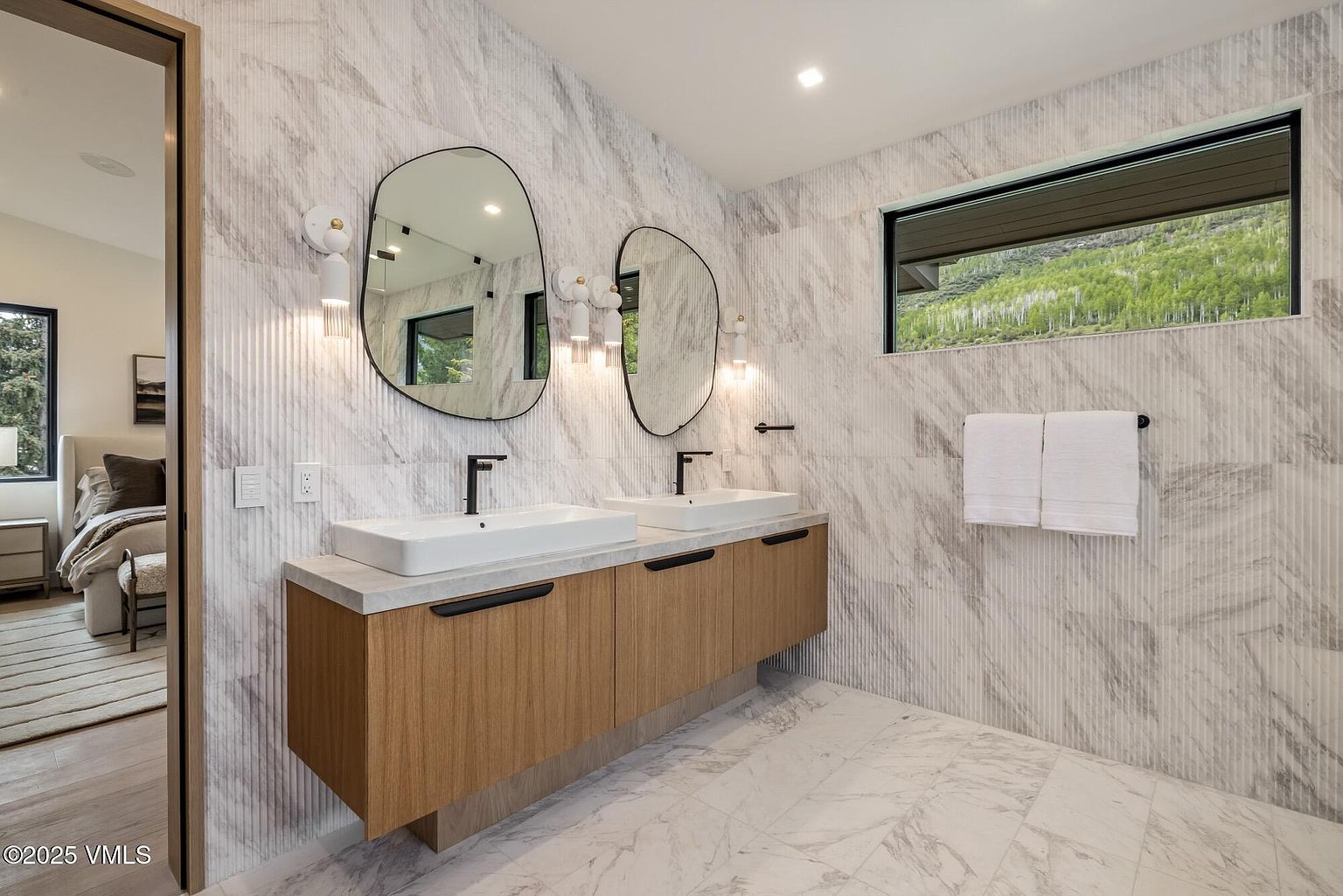 This modern primary bathroom features a floating dual-vanity with light wood cabinetry and a stone countertop, paired with two uniquely shaped mirrors. The walls are clad in vertically textured stone tiles, creating a sophisticated, spa-like atmosphere. A horizontal window above the towel rack provides natural light and a view of the outdoors, while the perspective captures the clean, contemporary design of the space.