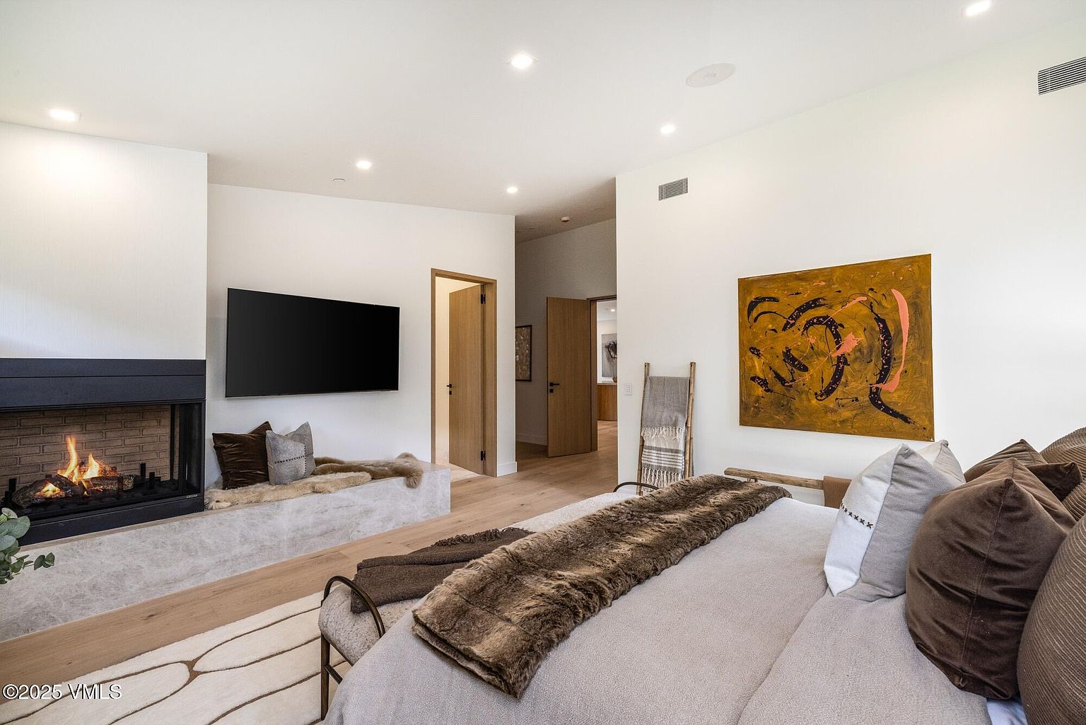 This primary bedroom features a cozy, modern aesthetic with a large bed dressed in neutral linens and a plush faux-fur throw. To the left, a stone-clad fireplace with a built-in bench provides a warm focal point, while a large abstract painting adds a vibrant pop of color to the wall. The room is finished with light wood flooring and recessed lighting, creating a serene and inviting atmosphere.