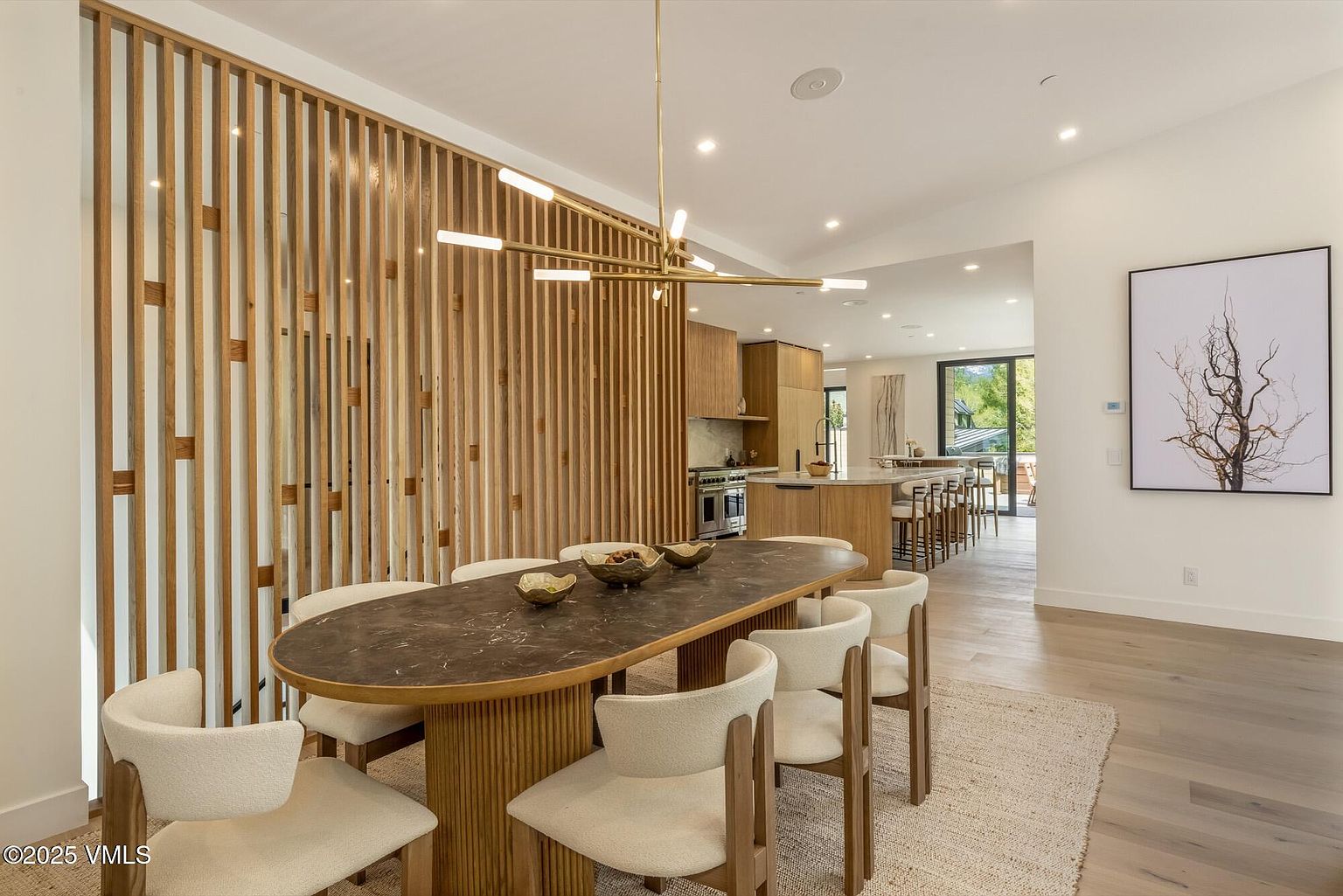 This elegant dining area features a dark, oval-shaped stone table surrounded by six upholstered chairs with light wood frames, set atop a textured area rug. A striking vertical wood slat screen serves as a room divider, separating the dining space from the adjacent kitchen, which is illuminated by a modern, multi-arm brass chandelier. The open-concept layout flows seamlessly into the kitchen, creating a bright and sophisticated atmosphere with light wood flooring and neutral walls.