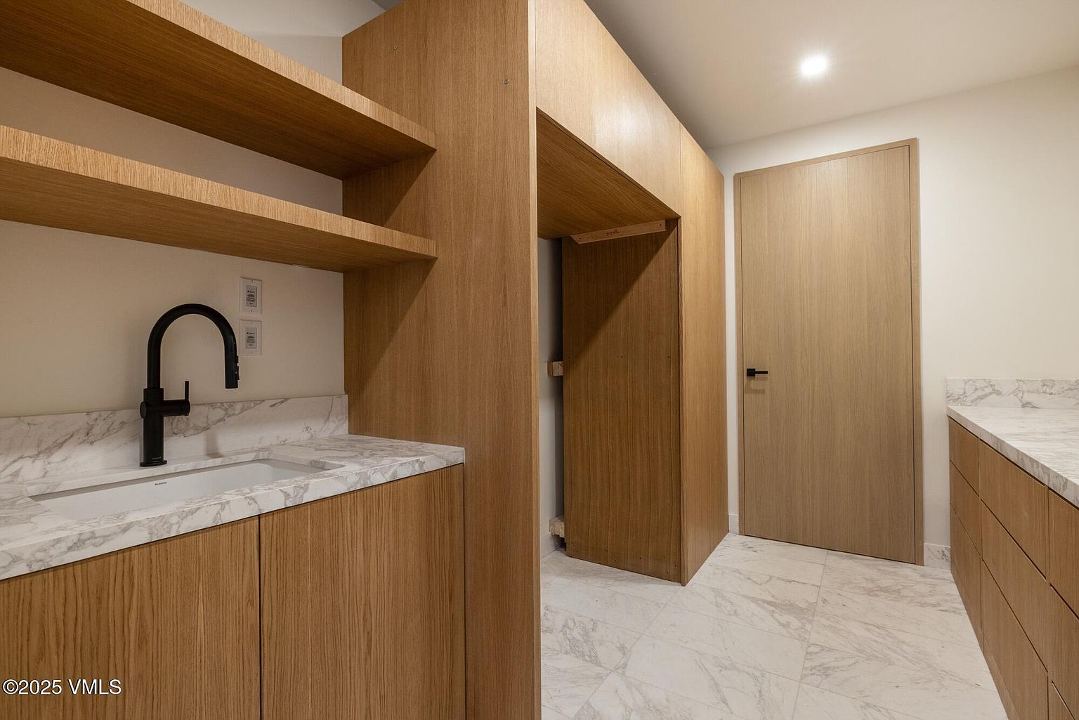 This modern laundry room features light wood cabinetry and a stone countertop with an integrated sink and a matte black faucet. The space includes open shelving, a dedicated alcove for appliances, and a matching light wood door, all set against a backdrop of light-toned stone flooring. The clean lines and neutral palette create a functional and contemporary aesthetic.