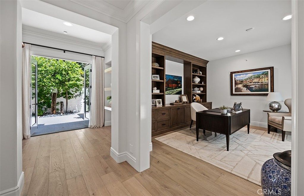 This elegant home office features light wood flooring and a sophisticated built-in desk and shelving unit with a dark finish. The room is bathed in natural light from a set of glass doors that open onto a private patio, creating a seamless indoor-outdoor connection. A patterned area rug anchors the workspace, while framed artwork and decorative accents add a refined, professional touch to the space.