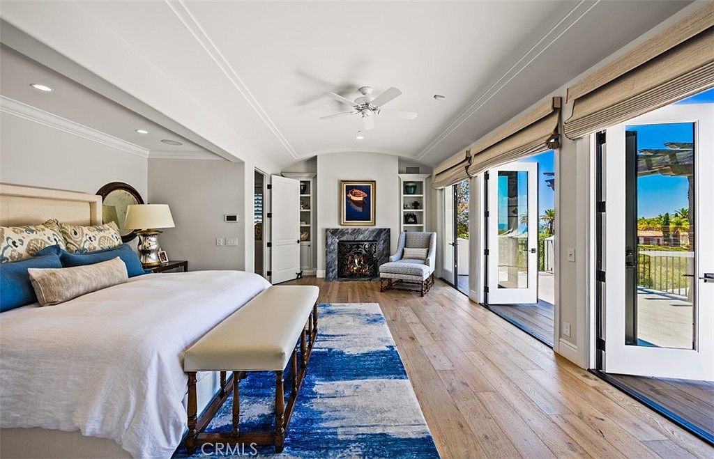This spacious primary bedroom features light wood flooring, a large bed with a neutral headboard, and a bench at the foot of the bed atop a blue and white patterned rug. A stone-clad fireplace serves as a focal point between built-in shelving units, while large glass doors open to an outdoor deck with scenic views. The room is bright and airy, characterized by a neutral color palette and a sophisticated, tranquil atmosphere.
