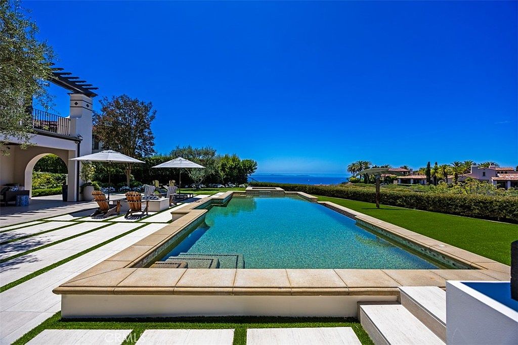 This expansive outdoor space features a rectangular swimming pool as the centerpiece, set against a backdrop of a clear blue sky and distant ocean views. The pool area is complemented by a manicured lawn, a paved patio with lounge seating under umbrellas, and a covered veranda attached to the main residence. The scene conveys a luxurious, resort-like atmosphere perfect for relaxation and entertaining.