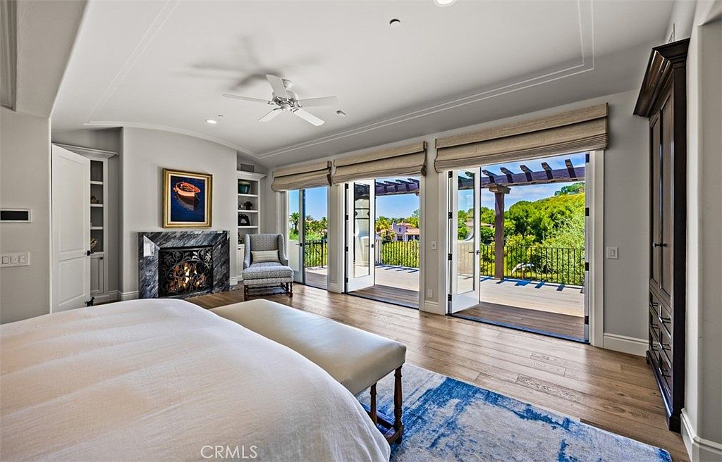 This spacious primary bedroom features light-toned wood flooring, a large bed with a neutral-colored bench at its foot, and a striking fireplace with a dark, patterned stone surround. Large glass doors open onto a private balcony, providing an expansive view of the surrounding hills and greenery. The room is accented by a patterned area rug, a comfortable armchair near the balcony, and built-in shelving, creating a serene and luxurious atmosphere.