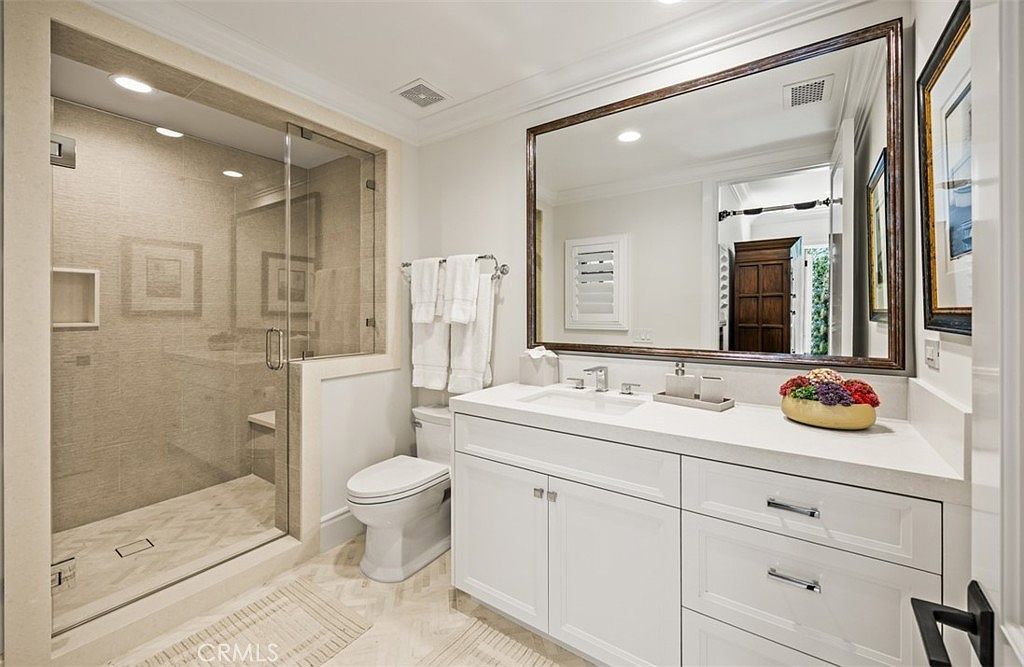 This bright and clean bathroom features a glass-enclosed walk-in shower with patterned tile flooring and a built-in bench. A white vanity with a stone countertop and a large, framed mirror provides a functional and elegant focal point. The space is finished with neutral tones and polished hardware, creating a polished and inviting atmosphere.