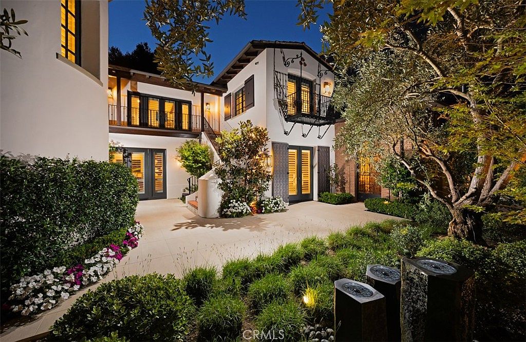 This elegant Mediterranean-style home features a multi-level exterior with white stucco walls, dark-framed windows, and a wrought-iron balcony. The paved courtyard is beautifully landscaped with lush greenery, flowering shrubs, and a trio of stone water features in the foreground. The scene is captured at twilight, with warm exterior lighting highlighting the architectural details and creating an inviting, sophisticated atmosphere.