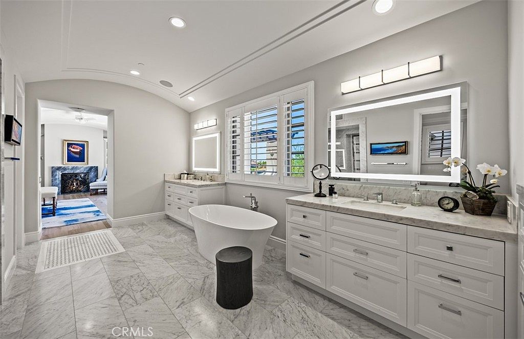 This spacious primary bathroom features a freestanding soaking tub as a central focal point, flanked by dual vanities with stone countertops and illuminated mirrors. The room is finished with light-toned walls, recessed lighting, and patterned stone flooring that extends into the adjacent room. A wide doorway provides a view into a secondary living space with a fireplace, creating an open and luxurious atmosphere.