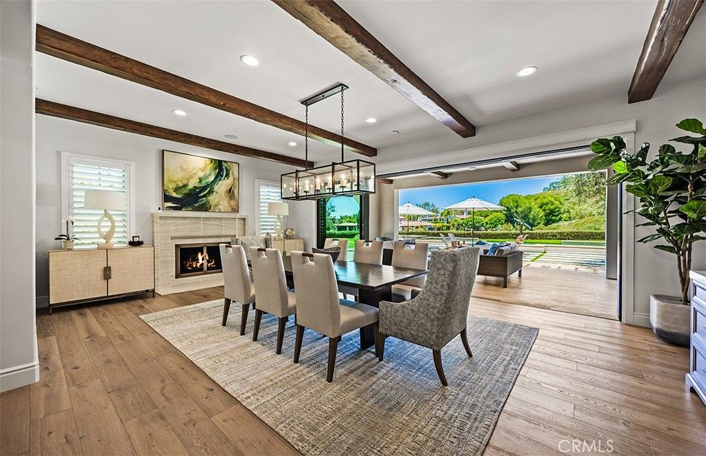 This elegant dining room features a large wooden table surrounded by upholstered chairs, set beneath a modern rectangular chandelier. The space is defined by exposed wooden ceiling beams and a light-colored stone fireplace surround, while a wide opening seamlessly connects the room to an expansive outdoor patio. The open-concept design and light wood flooring create a bright, airy atmosphere that emphasizes indoor-outdoor living.