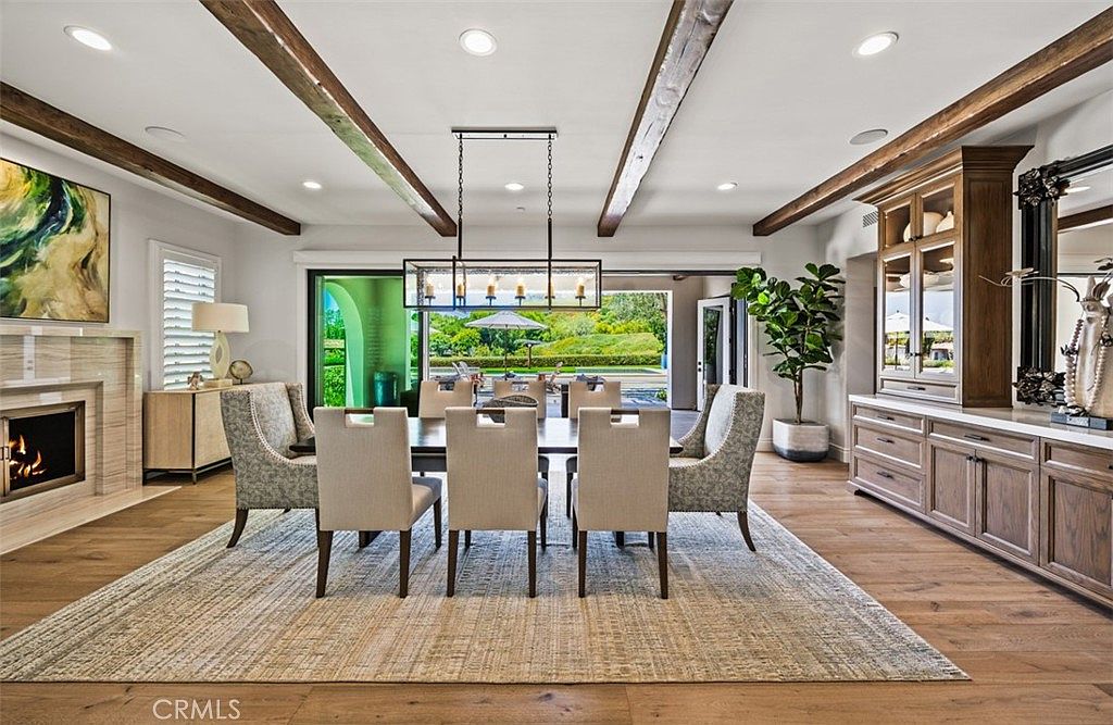 This elegant dining room features a large rectangular table surrounded by upholstered chairs, set beneath a modern linear chandelier. The space is defined by exposed wooden ceiling beams and light-toned wood flooring, with a tall stone-clad fireplace surround on the left and a built-in wooden hutch on the right. Large sliding glass doors open to a view of the outdoor patio and garden, creating a seamless transition between indoor and outdoor living.
