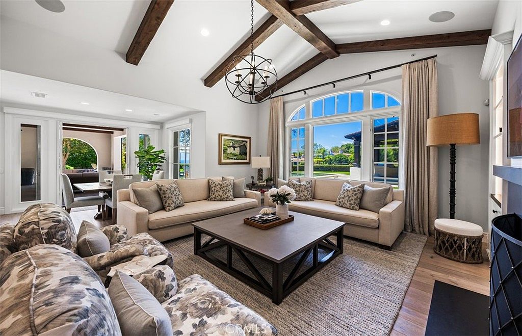 This spacious living room features a vaulted ceiling with exposed dark wood beams and a large, central chandelier. The room is furnished with two light-colored sofas facing a large, square coffee table, all set upon a textured area rug. Large, arched windows provide ample natural light and a view of the outdoor landscape, while a tall floor lamp stands near a fireplace on the right side of the frame.