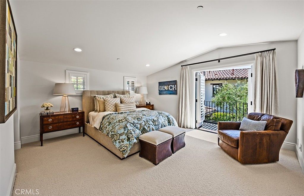 This spacious primary bedroom features a neutral color palette with a plush upholstered bed as the focal point, complemented by patterned bedding and two matching ottomans at the foot. To the right, a comfortable leather armchair sits near a set of French doors that open onto a private balcony, allowing for natural light to fill the room. The space is finished with a dark wood dresser, bedside lamps, and wall-to-wall carpeting, creating a serene and inviting atmosphere.
