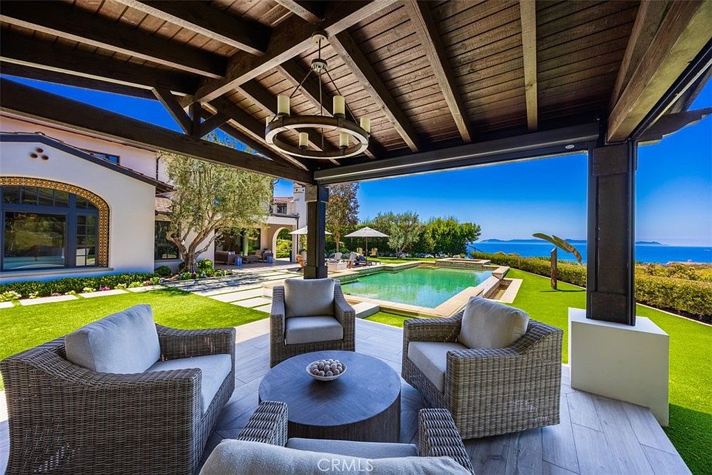 This luxurious covered patio features a comfortable outdoor seating area with wicker furniture arranged around a circular coffee table, all sheltered by a rustic wood-beamed pergola. The space opens directly onto a manicured lawn and a sparkling swimming pool, with a stunning, unobstructed panoramic view of the ocean and distant islands in the background. The combination of high-end outdoor living space and breathtaking coastal scenery creates an inviting and serene atmosphere.
