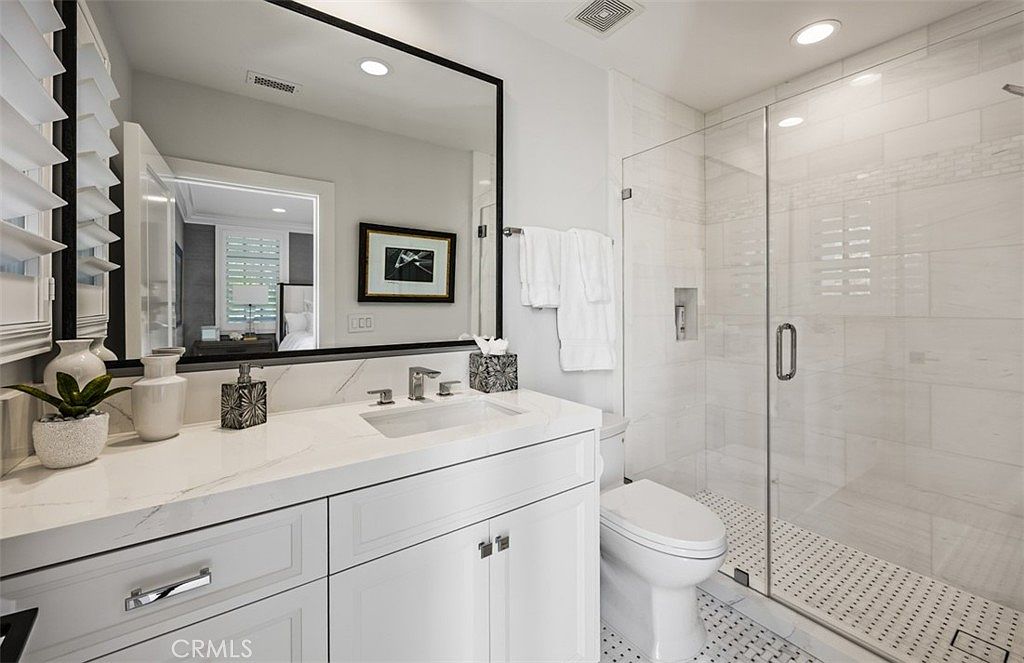 This bright and modern bathroom features a white vanity with a stone countertop, a large rectangular mirror with a black frame, and a glass-enclosed walk-in shower. The shower is finished with light-colored wall tiles and a patterned tile floor, creating a clean and cohesive aesthetic. The space is well-lit by recessed ceiling lights and includes a white toilet positioned next to the shower.
