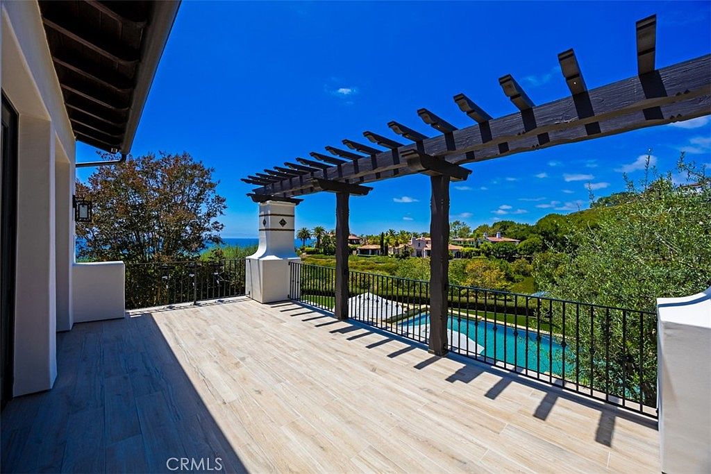 This elevated balcony features light wood-look tile flooring and a dark wood pergola that provides partial shade. A black metal railing borders the space, offering an unobstructed view of the lush green landscape, a swimming pool below, and the distant ocean horizon. The perspective is captured from the balcony looking outward, emphasizing the seamless transition between indoor living and the scenic outdoor environment.