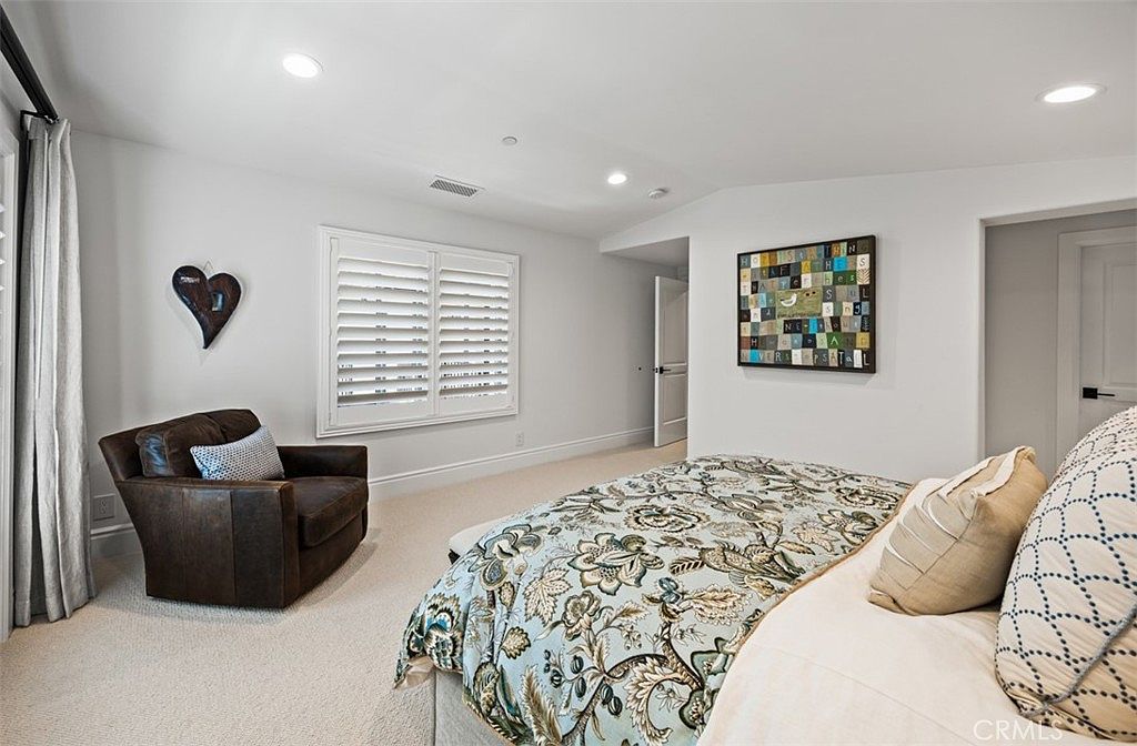 This bright and airy bedroom features a neutral color palette with white walls and light-toned carpeting. The room is furnished with a comfortable leather armchair near a window with plantation shutters and a bed dressed in a patterned floral duvet. A decorative wall art piece adds a touch of personality to the space, which is illuminated by recessed lighting.