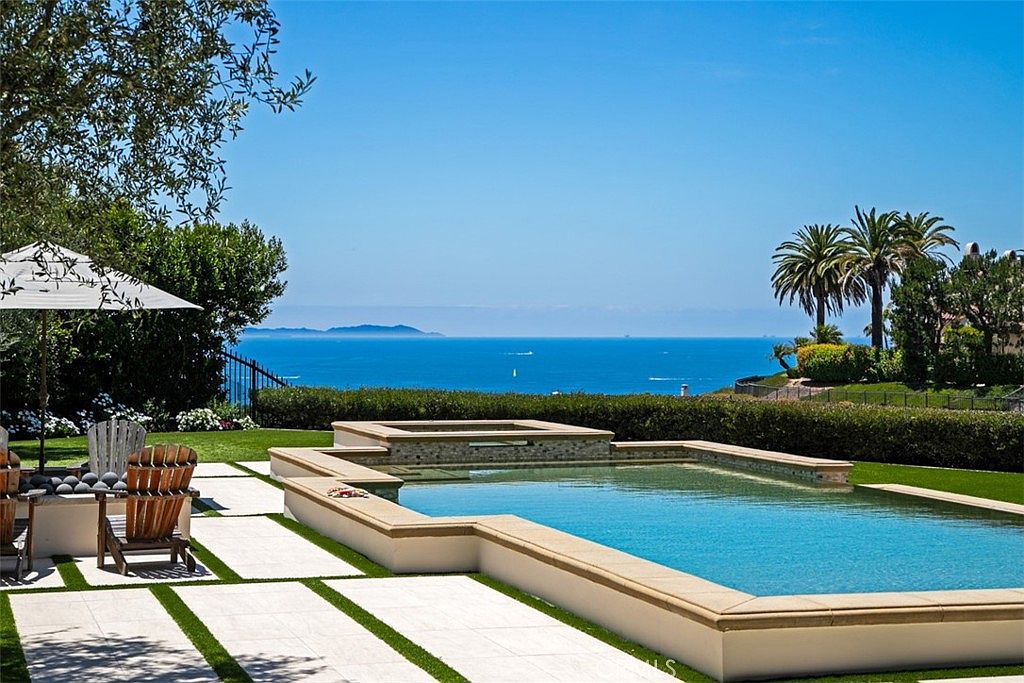 This stunning outdoor space features a raised infinity-edge pool and integrated spa overlooking a vast ocean horizon. The patio area is paved with large, light-colored stone tiles set in a grid pattern with grass inlays, complemented by wooden lounge chairs and a white umbrella. The scene is framed by lush green hedges and mature palm trees, creating a serene and luxurious coastal atmosphere.