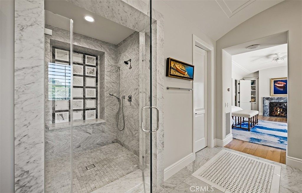 This primary bathroom features a spacious walk-in shower enclosed by a glass door, showcasing light-toned stone wall tiles and a grid-patterned window. A white door leads to an adjacent room, while a patterned tile rug sits on the floor in the foreground. Through the doorway, a glimpse of a bedroom with a fireplace and a blue area rug creates a sense of depth and continuity between the spaces.