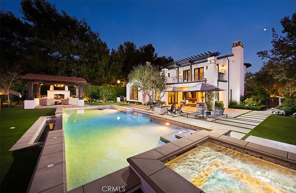 This twilight view captures a luxurious backyard oasis featuring a large, illuminated swimming pool and a foreground spa. The two-story white stucco residence serves as a stunning backdrop, complete with a second-story balcony, a pergola-covered patio area, and a separate outdoor pavilion with a fireplace. The scene is professionally landscaped with manicured lawns and mature trees, creating an inviting and sophisticated atmosphere for outdoor entertaining.