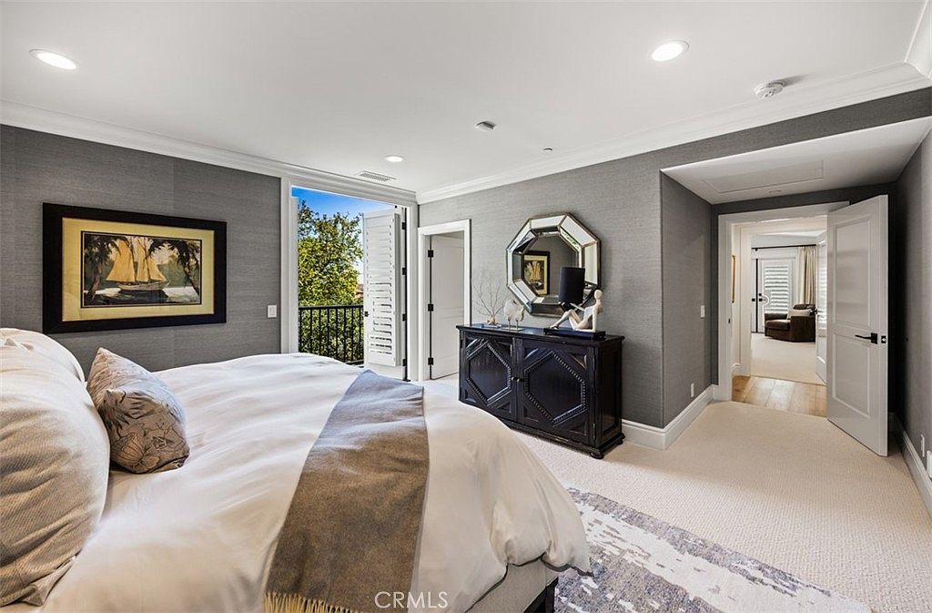 This elegant primary bedroom features textured gray wallpaper, plush light-colored carpeting, and a large bed dressed in neutral linens. A dark wood dresser topped with a decorative mirror and sculpture serves as a focal point, while a doorway leads to an adjacent room. The space includes a balcony with a wrought-iron railing, allowing natural light to fill the room through open shutters.