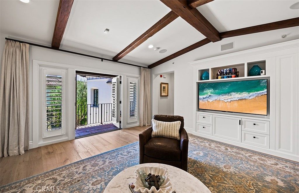 This elegant living room features exposed dark wood ceiling beams and light-toned wood flooring, creating a warm and sophisticated atmosphere. A large, built-in white media cabinet serves as a focal point, housing a television and decorative accents, while a leather armchair sits atop a detailed patterned rug. The room opens through double doors onto a small balcony, allowing natural light to flood the space and providing a seamless transition to the outdoors.