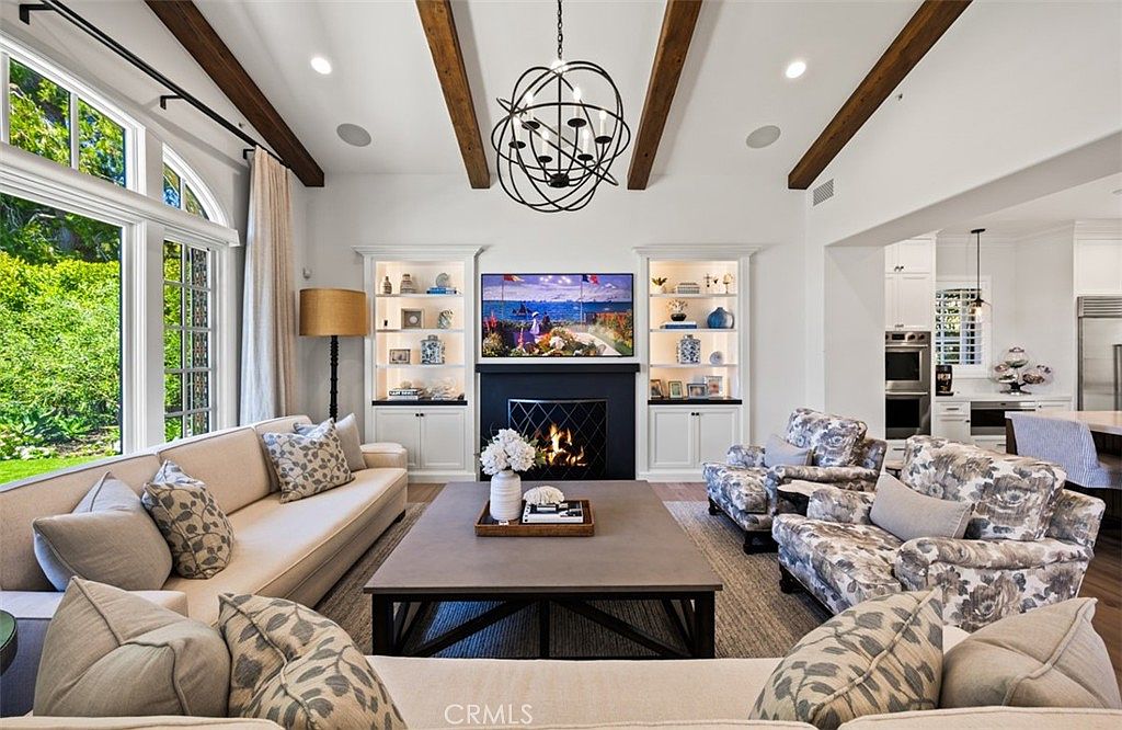 This bright and airy living room features a vaulted ceiling with exposed wooden beams and a large, modern spherical chandelier. A central fireplace with a dark surround serves as the focal point, flanked by built-in white shelving units. The space is furnished with a large neutral sofa and patterned armchairs, all arranged around a substantial rectangular coffee table on a textured area rug.