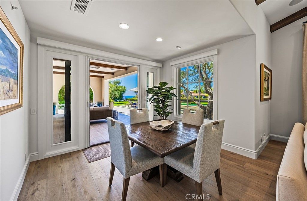 This bright dining area features a dark wood square table surrounded by four upholstered chairs, creating an intimate setting. Large glass doors open to a covered patio, seamlessly blending the indoor space with the outdoor landscape and ocean view. The room is finished with light wood flooring and neutral walls, providing a clean and airy atmosphere.