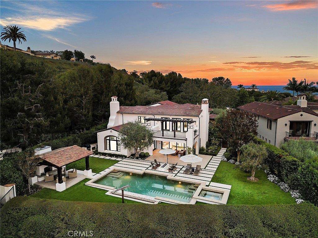 This aerial perspective captures a luxurious Mediterranean-style residence at sunset, showcasing a beautifully landscaped backyard with a swimming pool and a covered outdoor lounge area. The home features a light-colored stucco exterior, a tiled roof, and multiple balconies overlooking the manicured lawn and surrounding greenery. The scene is bathed in the warm, golden light of dusk, highlighting the property's elegant architecture and serene outdoor living spaces.