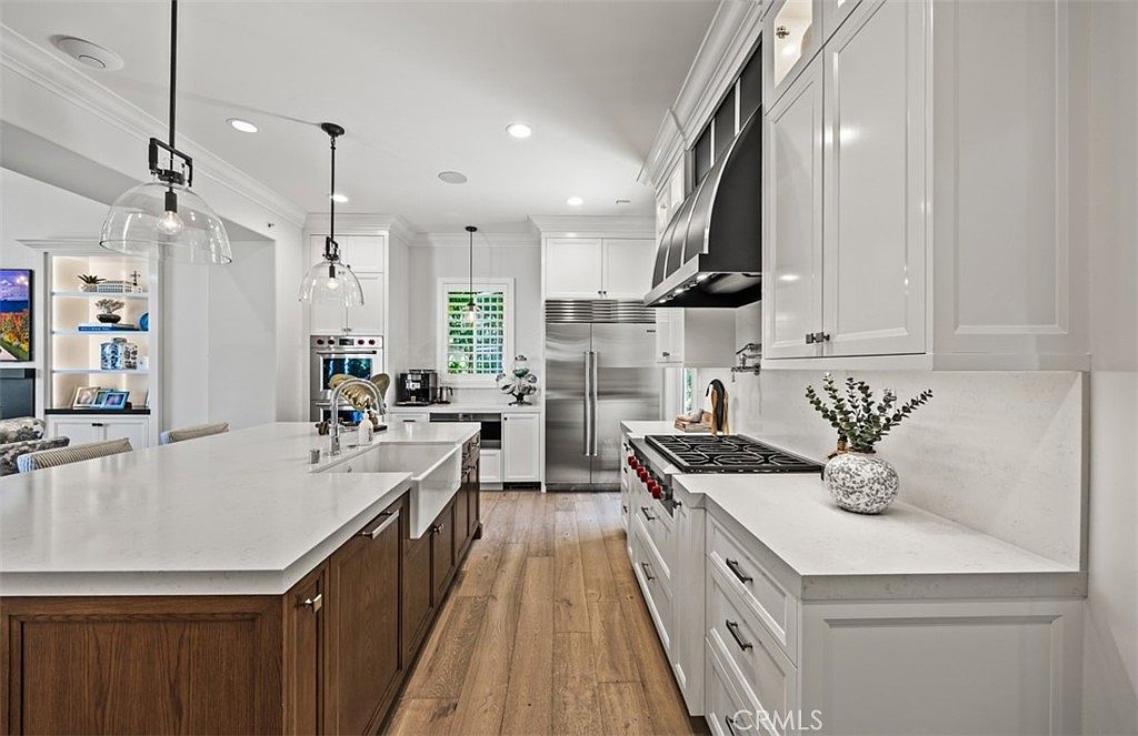 This spacious, high-end kitchen features a large central island with a farmhouse sink and dark wood cabinetry, contrasting with the bright white perimeter cabinets and stone countertops. A professional-grade range with a prominent black hood serves as a focal point, complemented by stainless steel appliances and warm wood flooring. The perspective is a wide-angle shot from the kitchen entrance, showcasing an open-concept layout that flows into an adjacent living area with built-in shelving.