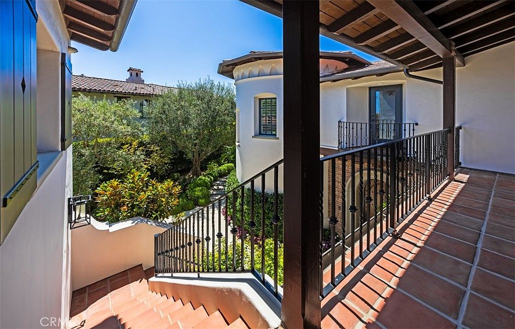 This image captures a Mediterranean-style outdoor balcony and staircase area, featuring terracotta-tiled flooring and dark wrought-iron railings. The perspective looks down from an upper-level landing, showcasing a lush, sun-drenched courtyard garden with mature trees and manicured greenery below. The architectural design includes a distinctive rounded tower structure, creating a sophisticated and inviting atmosphere typical of high-end residential estates.