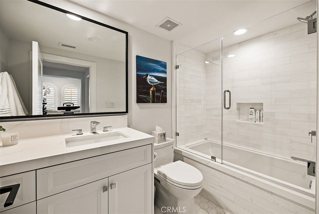 This bright and modern bathroom features a white vanity with a stone countertop and a large, black-framed mirror. To the right, a glass-enclosed shower and tub combination is finished with light-colored subway tile and includes a built-in recessed niche for storage. The space is accented by a piece of wall art depicting a bird, creating a clean and contemporary aesthetic.