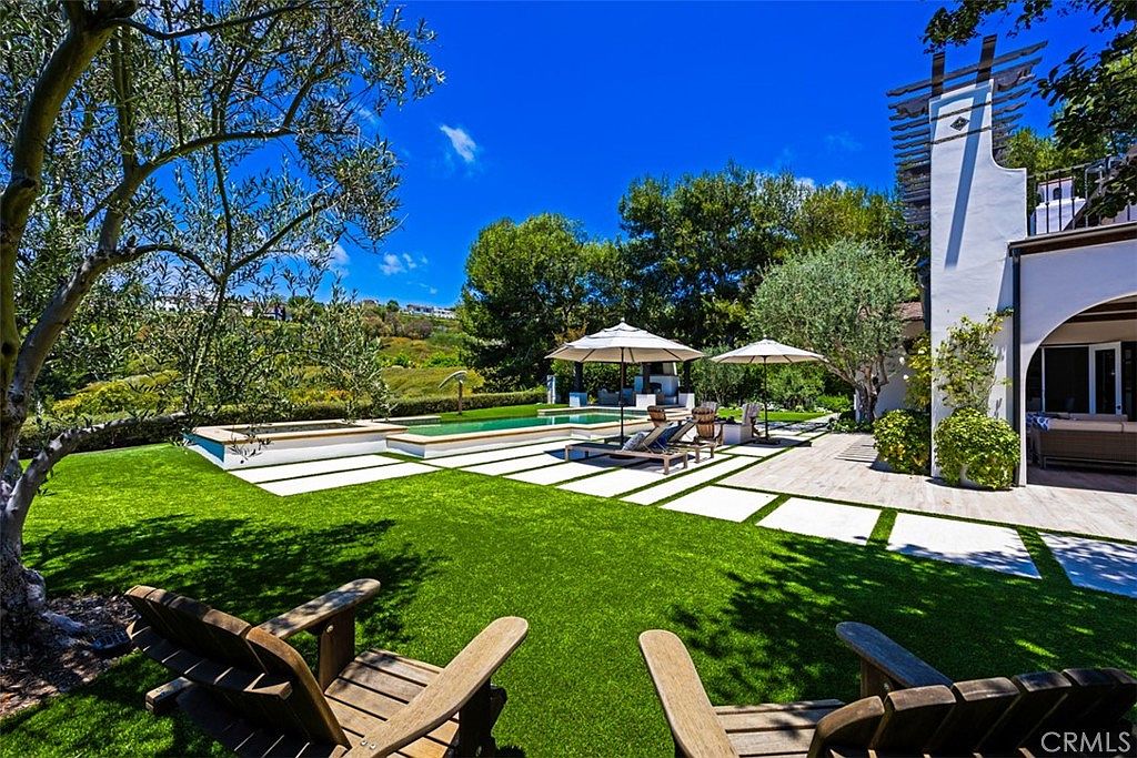 This expansive backyard features a lush green lawn accented by a geometric pattern of large stone pavers leading toward a rectangular swimming pool. The scene includes two wooden lounge chairs in the foreground, a pair of patio umbrellas, and a white stucco structure with a pergola-style roof detail. The perspective is a wide-angle ground-level shot that captures the serene, resort-like atmosphere of the outdoor living space.