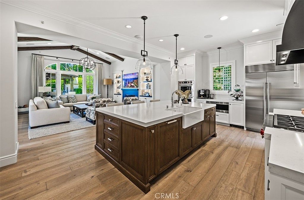 This open-concept kitchen features a large central island with a farmhouse sink and dark wood cabinetry, contrasting with the surrounding white perimeter cabinets. The space flows seamlessly into a bright living area with exposed ceiling beams and large windows that provide a view of the outdoors. The overall aesthetic is sophisticated and airy, highlighting a blend of traditional warmth and modern design elements.