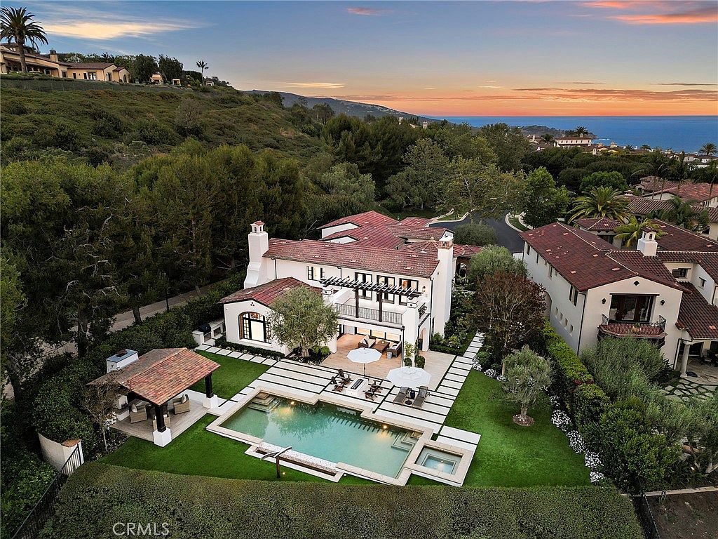 This high-angle aerial view captures a luxurious Mediterranean-style estate at sunset, overlooking a coastal landscape. The property features a multi-level residence with a red-tiled roof, a private swimming pool with an integrated spa, and a covered outdoor pavilion set within a manicured lawn. The scene is framed by lush greenery and mature trees, emphasizing the home's secluded and upscale atmosphere.