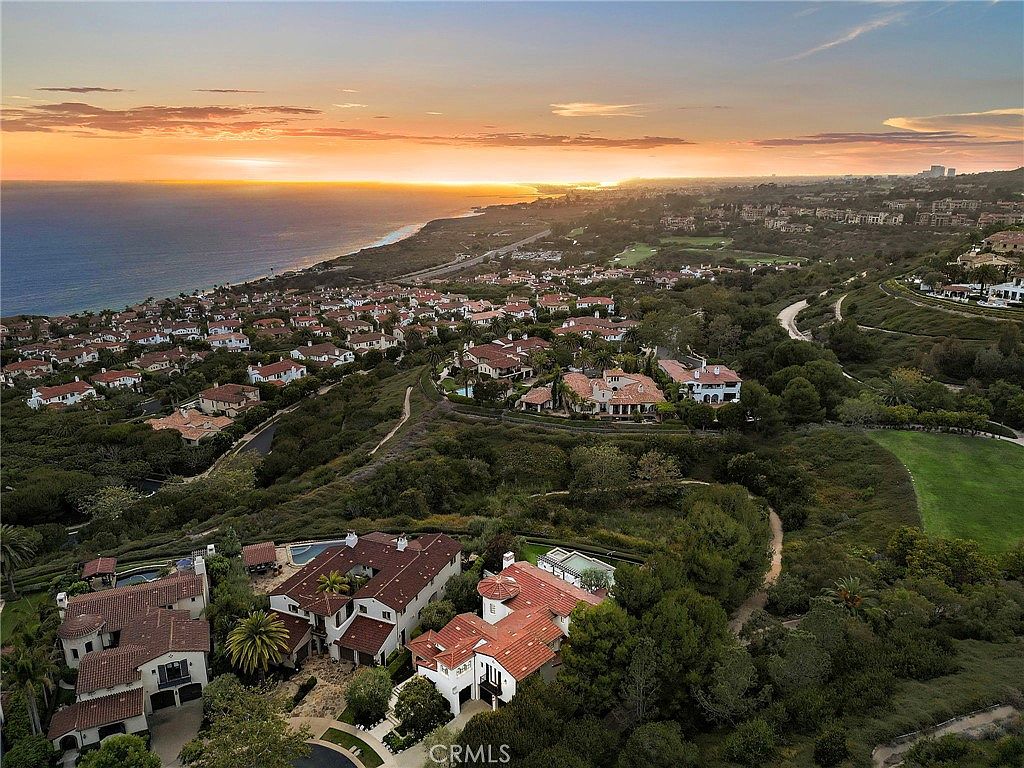 This high-angle aerial view captures a sprawling coastal residential community at sunset, showcasing Mediterranean-style homes with terracotta-tiled roofs. The perspective highlights the proximity of the properties to the ocean, with a winding road and lush green landscaping weaving through the neighborhood. The warm, golden light of the setting sun reflects off the water and illuminates the hillside, creating a serene and luxurious atmosphere.