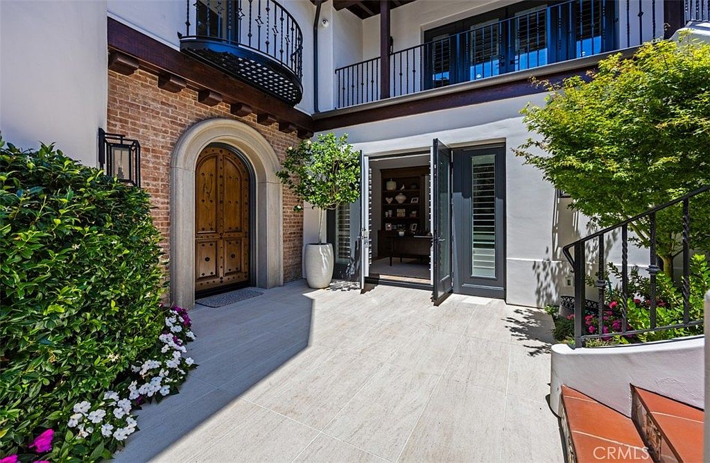 This elegant courtyard entryway features a rustic arched wooden door set into a brick-accented wall, complemented by a modern dark-framed glass door leading into an interior office space. The area is paved with light-toned stone tiles and includes a potted tree, lush greenery, and a wrought-iron staircase leading to an upper balcony. The architectural style blends Mediterranean influences with contemporary design elements, creating a sophisticated and inviting transition space.