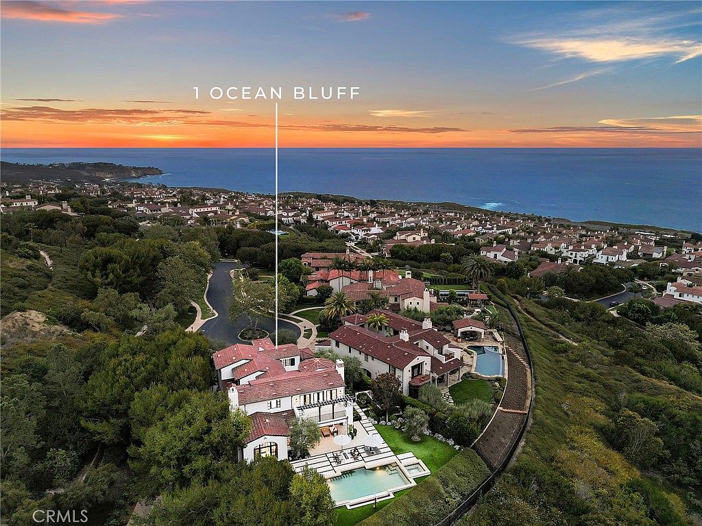 This high-angle aerial view captures a sprawling Mediterranean-style estate perched on a coastal bluff overlooking the ocean. The property features a multi-level residence with a red-tiled roof, a private swimming pool, and manicured landscaping, all set against a dramatic sunset horizon. The perspective emphasizes the home's exclusive location within a dense, upscale residential community.