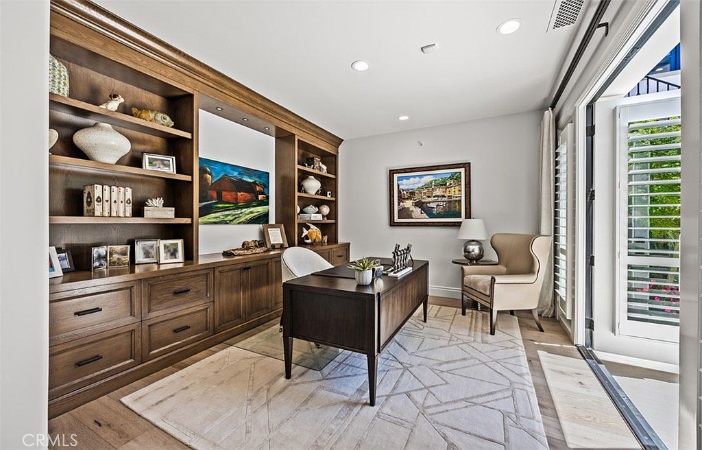 This sophisticated home office features extensive built-in dark wood cabinetry and shelving that frame a central desk. A geometric patterned area rug anchors the space, while a comfortable wingback chair sits near a set of glass doors that open to the outdoors. The room is well-lit with recessed lighting and offers a professional yet inviting atmosphere.