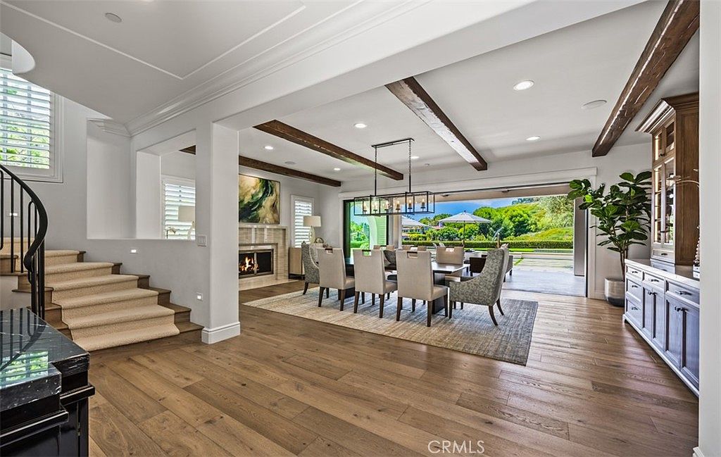 This elegant dining room features an open-concept layout with exposed wooden ceiling beams and light-toned wood flooring. A large dining table is centered under a modern linear chandelier, flanked by upholstered chairs, while a fireplace with a stone surround adds warmth to the adjacent wall. The space seamlessly transitions to an outdoor patio through expansive sliding glass doors, creating a bright and airy atmosphere perfect for entertaining.