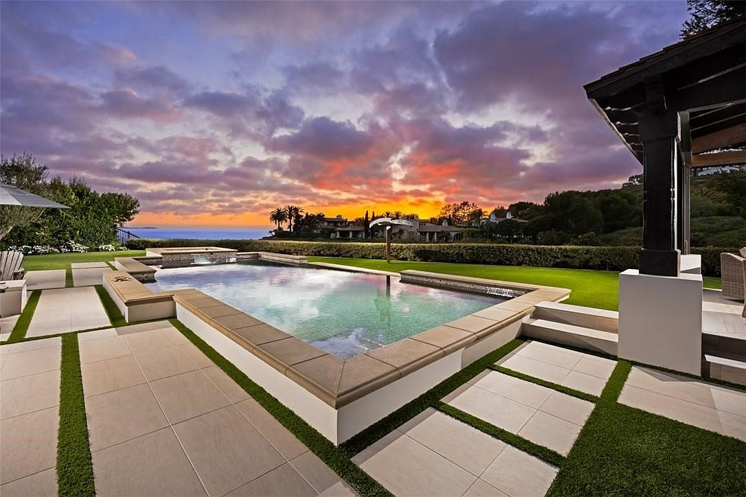 This stunning backyard features a rectangular swimming pool with a raised spa and integrated water feature, set against a dramatic sunset sky. The surrounding patio is composed of large, light-toned pavers interspersed with strips of manicured artificial turf, creating a clean, modern aesthetic. A covered patio structure with dark wooden beams provides a shaded transition area, while the distant horizon offers a glimpse of the ocean beyond the landscaped grounds.