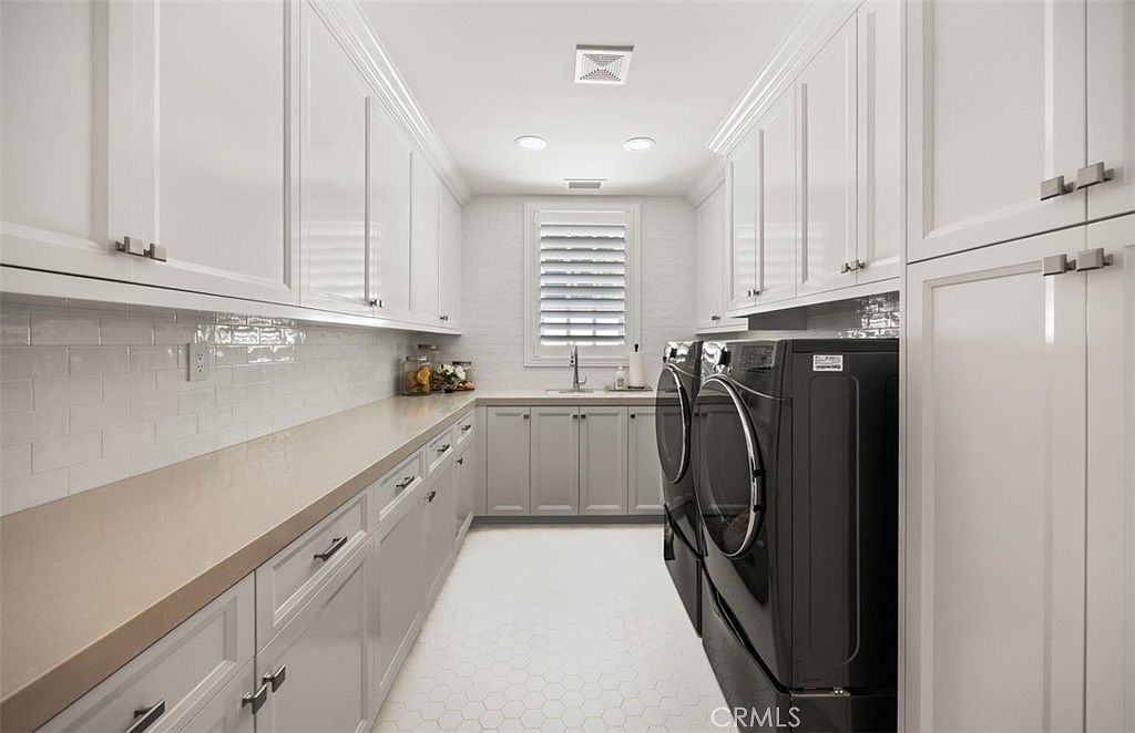 This bright and organized laundry room features crisp white upper cabinetry and light gray lower cabinets topped with a neutral stone countertop. The space includes a deep undermount sink, a white subway tile backsplash, and a pair of black front-loading laundry appliances. A window with white shutters provides natural light, while the floor is finished with a clean, white hexagonal tile pattern.