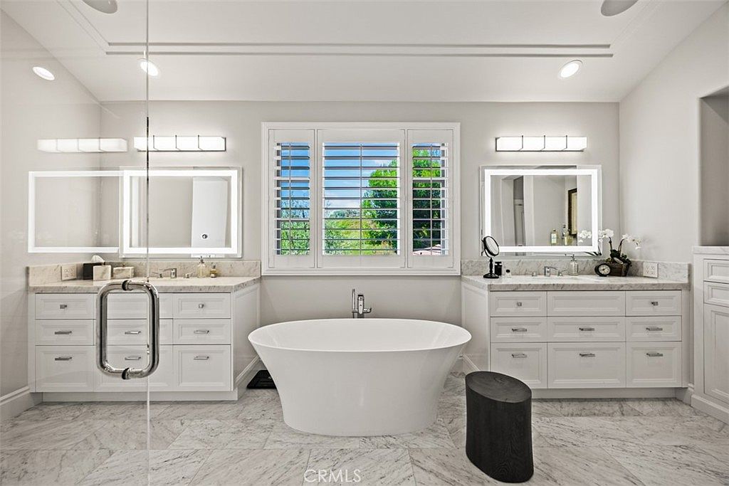 This luxurious primary bathroom features a symmetrical layout with dual vanities flanking a central freestanding soaking tub positioned beneath a large window. The space is finished with light-toned cabinetry, stone countertops, and patterned tile flooring, creating a bright and clean aesthetic. Modern illuminated mirrors and sleek light fixtures enhance the contemporary design, while a glass-enclosed shower area is visible on the left.