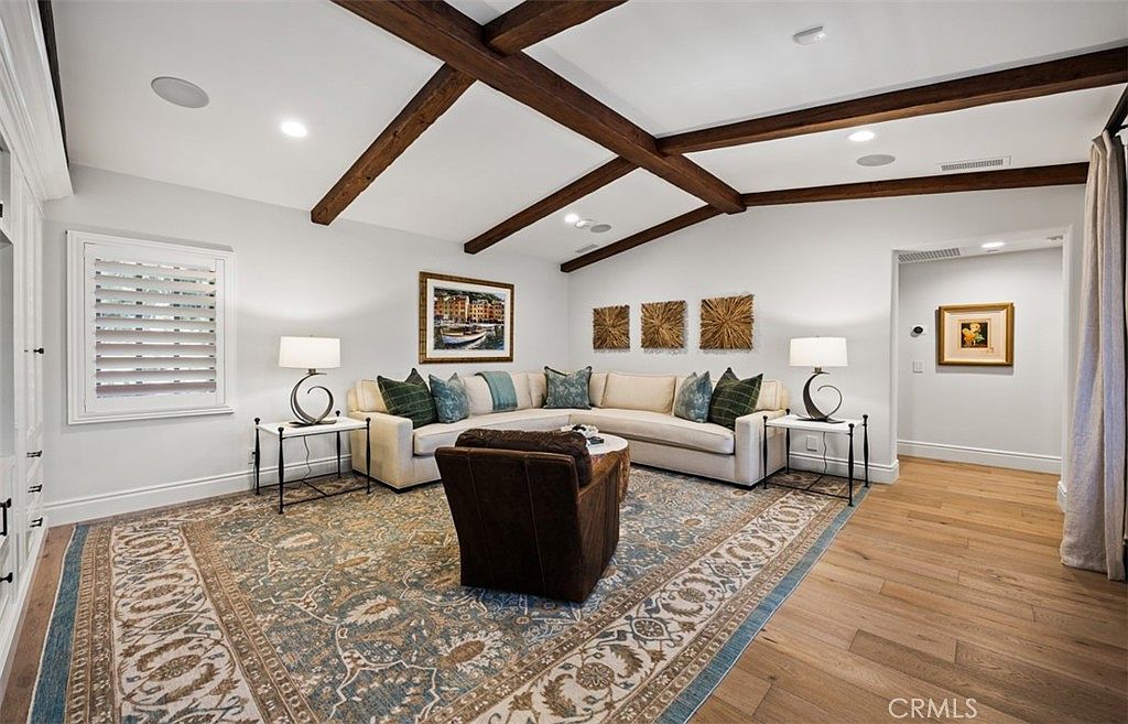 This inviting living room features a large, neutral-toned sectional sofa adorned with patterned accent pillows, centered around a circular coffee table and a dark leather armchair. The space is anchored by a large, intricately patterned area rug over light wood flooring, while exposed dark wood ceiling beams add architectural character. The room is illuminated by two matching table lamps on side tables and recessed lighting, creating a warm and comfortable atmosphere.