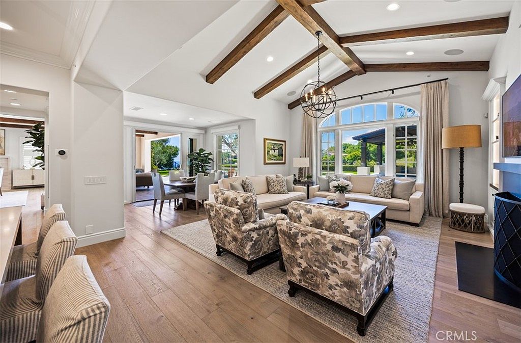 This spacious living room features a vaulted ceiling with exposed wooden beams and a large, multi-pane window arrangement that floods the space with natural light. The room is furnished with two light-colored sofas and two patterned armchairs arranged around a central coffee table, all resting on a large area rug. A dining area is visible in the background, creating an open-concept flow that leads toward an outdoor patio space.