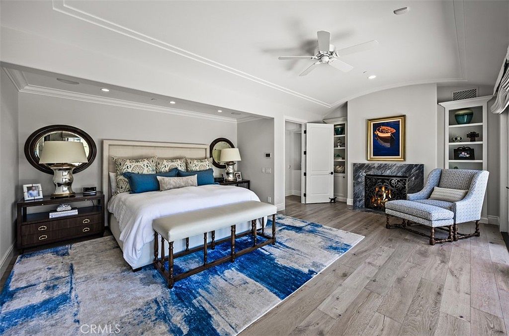 This spacious primary bedroom features a neutral color palette accented by a bold, abstract blue and white area rug. The room includes a large bed with a tufted headboard, a matching bench at the foot, and a cozy seating area beside a stone-clad fireplace. Light wood flooring and a ceiling fan complete the elegant, well-lit space.