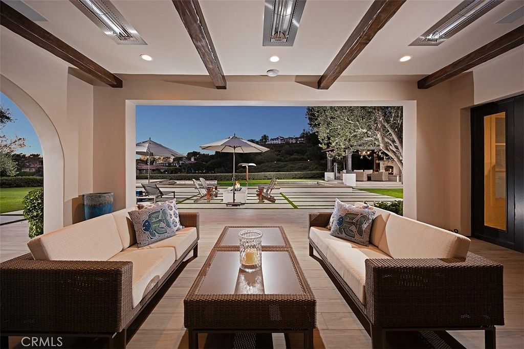 This covered outdoor living space features a symmetrical arrangement of two wicker sofas facing a central coffee table, creating an inviting lounge area. The space opens directly to a landscaped backyard with a pool, fire pit, and lounge chairs, framed by a large, unobstructed opening. Overhead, exposed wooden beams and recessed lighting add warmth to the structure, while the neutral color palette and clean lines emphasize a sophisticated, resort-style aesthetic.