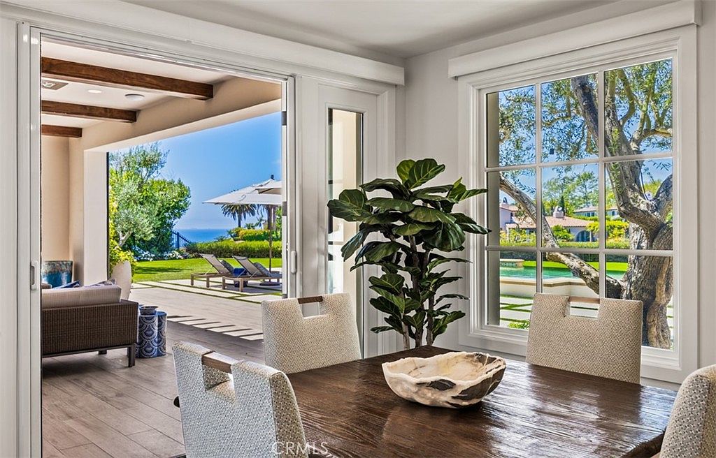 This bright dining area features a dark wood table centered in the room, surrounded by light-colored upholstered chairs. Large sliding glass doors open to a patio with a scenic ocean view, while a window on the right provides a view of a lush garden and pool area. A potted plant adds a touch of greenery to the space, creating an airy and inviting atmosphere that seamlessly blends indoor and outdoor living.