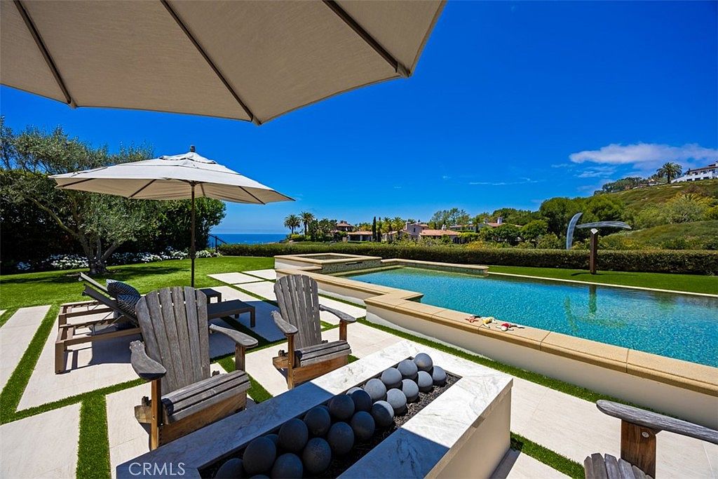 This luxurious outdoor living space features a rectangular swimming pool and an adjacent spa, set against a backdrop of rolling hills and a distant ocean view. The foreground includes a stone-surfaced patio area with wooden Adirondack chairs arranged around a fire pit filled with decorative dark spheres, all shaded by a large umbrella. The manicured lawn and lush landscaping create a serene, resort-like atmosphere perfect for relaxation and entertaining.