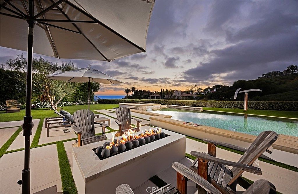 This luxurious outdoor living space features a rectangular fire pit with dark decorative spheres, surrounded by wooden Adirondack chairs on a paved patio. A sleek, infinity-edge pool sits adjacent to the seating area, overlooking a manicured lawn and a distant coastal horizon under a dramatic, cloud-filled sunset sky. Large umbrellas provide shade, creating an inviting atmosphere for evening relaxation and entertaining.