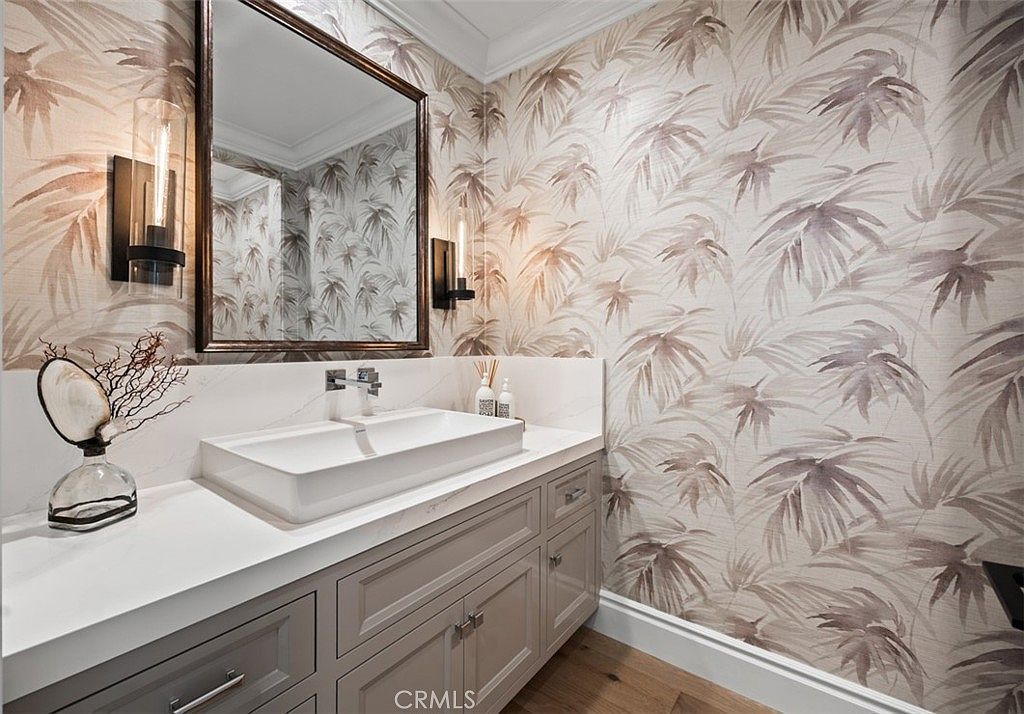 This elegant powder room features striking palm-leaf patterned wallpaper and a sleek, rectangular vessel sink set atop a light-toned stone vanity. The space is accented by a dark-framed mirror and modern wall-mounted light fixtures, creating a sophisticated and polished aesthetic. The vanity includes ample storage with light-colored cabinetry and brushed metal hardware, complemented by warm-toned wood flooring.