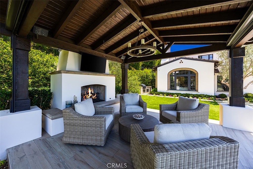 This covered outdoor living space features a rustic wood-beamed ceiling, a central chandelier, and a white fireplace with a mounted television. The patio is furnished with comfortable wicker seating arranged around a circular coffee table, overlooking a manicured lawn and the main house. The space offers a seamless transition between indoor comfort and outdoor relaxation, perfect for entertaining.